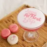 A pink cocktail and pink macarons printed with Valentine's Day designs. The cocktail says Love at first sip